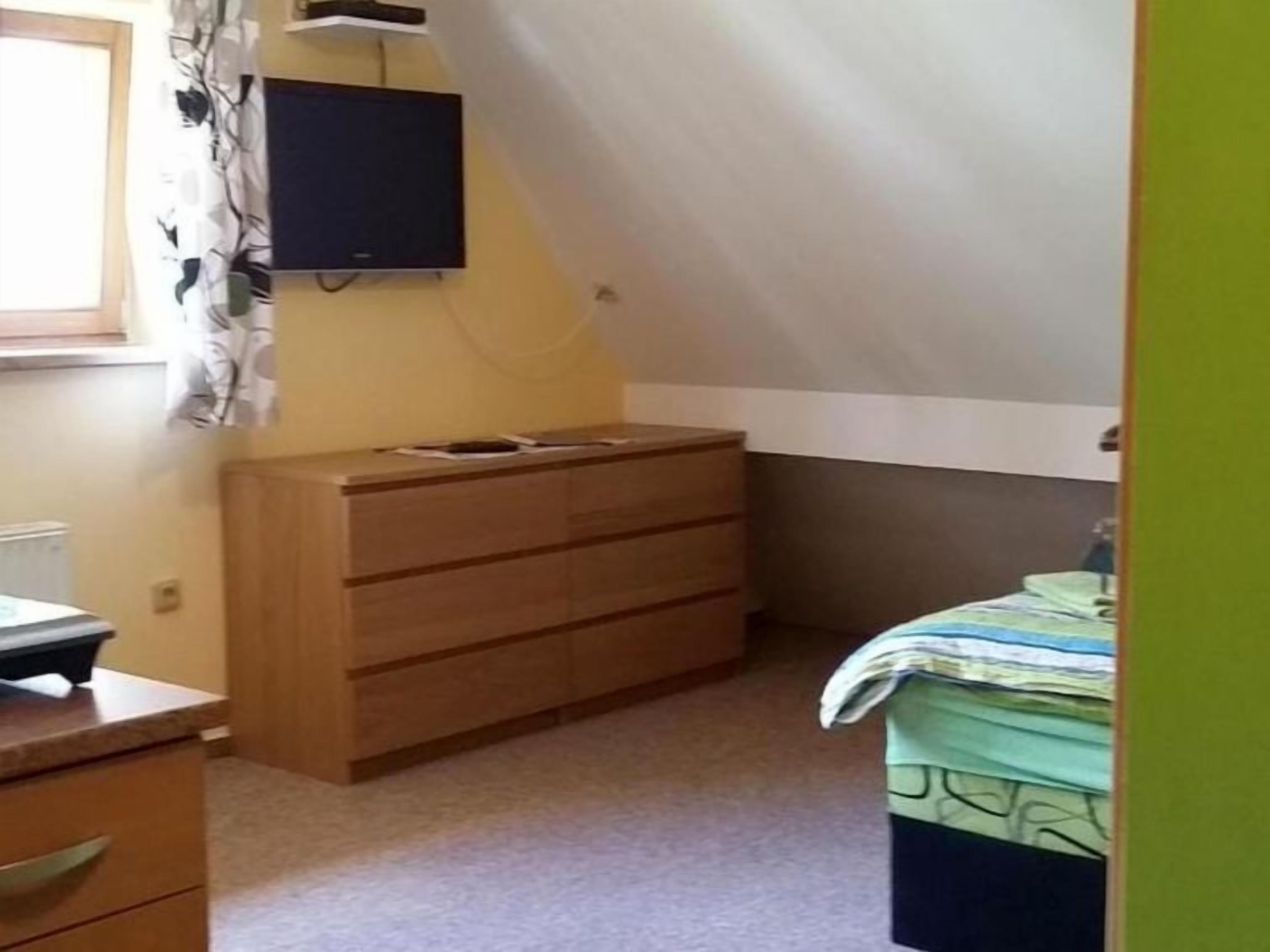 Ein Schlafzimmer mit einem hölzernen Schminktisch, einem Bett mit einer grün-weißen Tagesdecke, einem an der Wand befestigten Fernseher und einem Fenster mit Vorhängen.