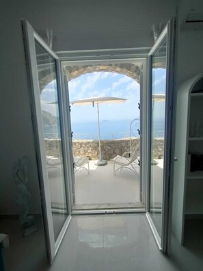 A room with a glass door leading to an outdoor patio with a white umbrella and two white chairs. - Positanobay (Positano)