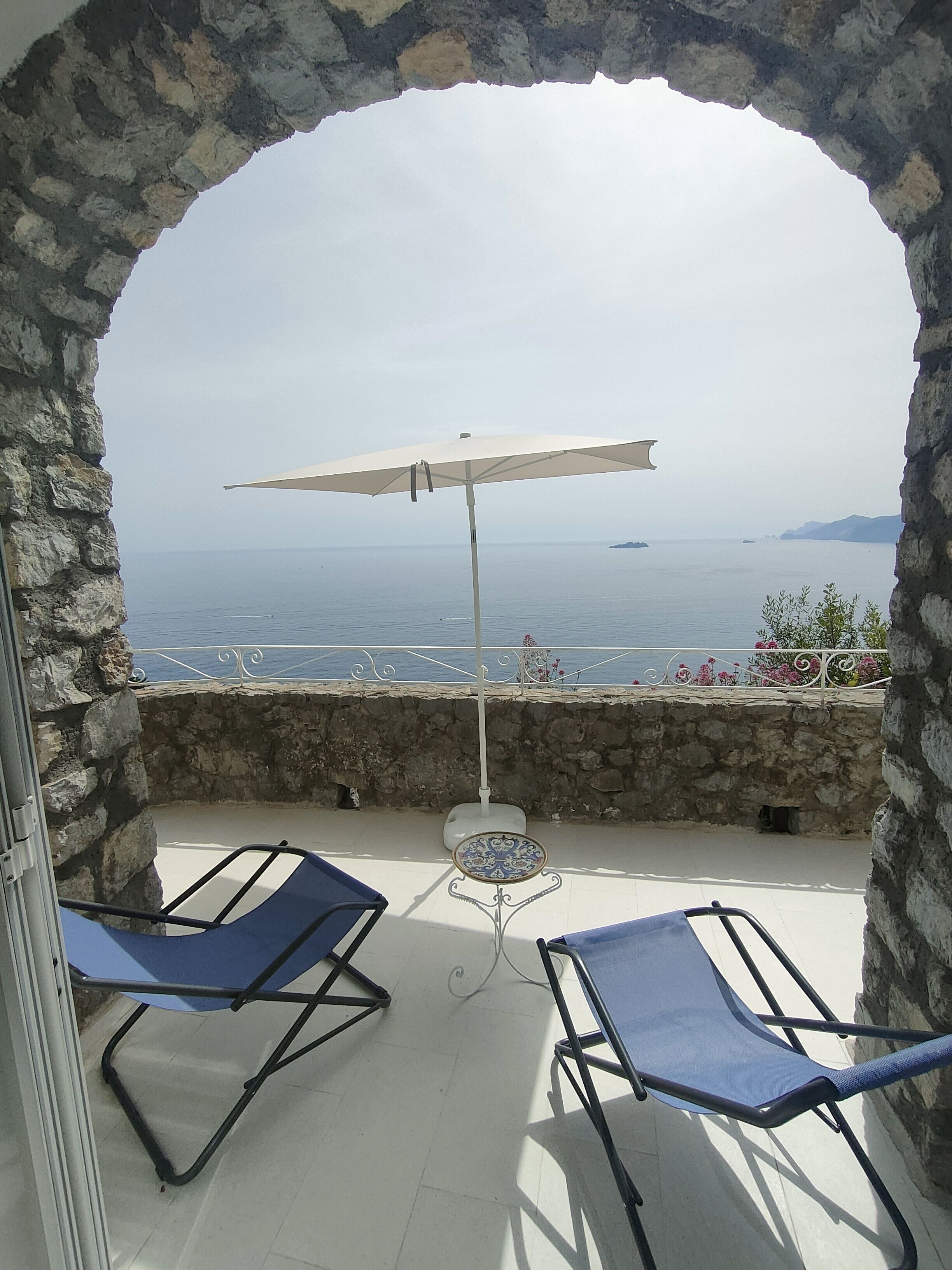Un balcon avec deux fauteuils bleus et une table recouverte d’une nappe à motifs, offrant une vue sur la mer.