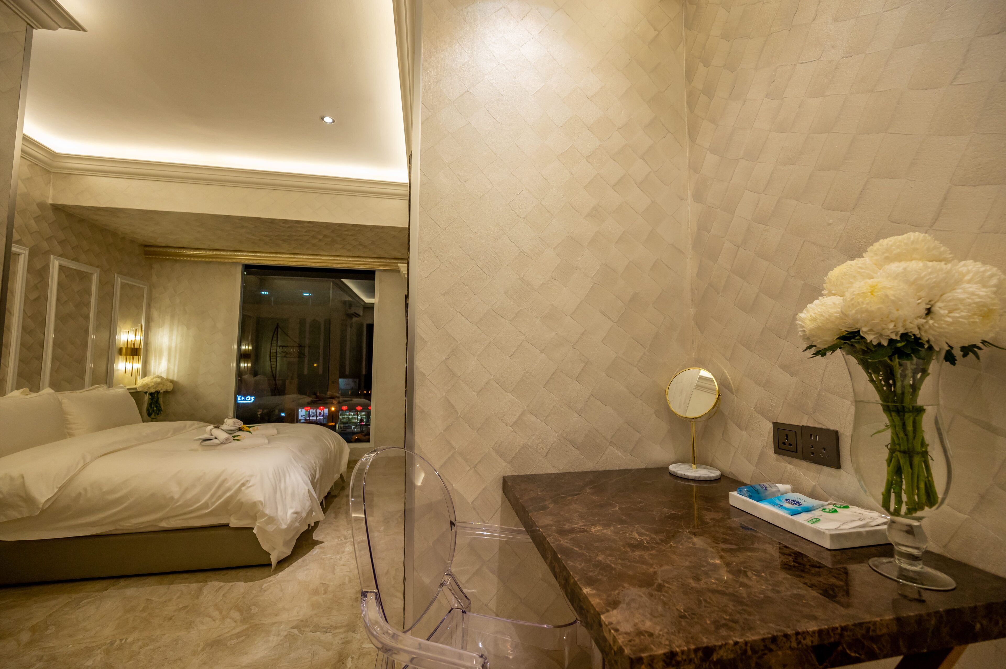 A hotel room with a bed, a bathroom vanity, a vase with flowers, and a night view through the window.