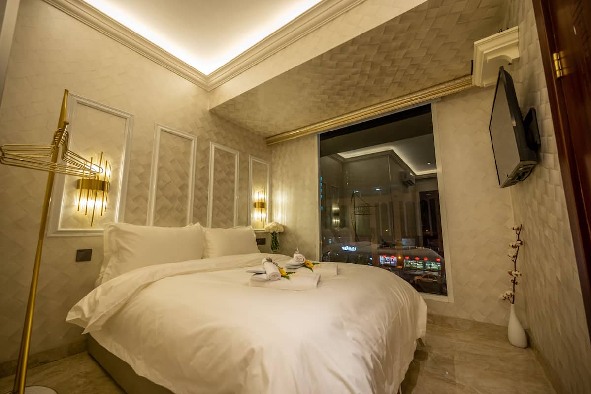 A hotel room with a large bed, a floor lamp, a TV mounted on the wall, and a window offering a view of the city at night.