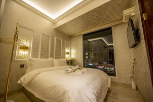A hotel room with a large bed, a floor lamp, a TV mounted on the wall, and a window offering a view of the city at night.