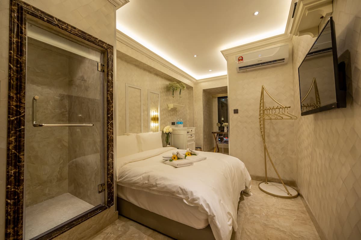 A modern hotel room with a marble shower enclosure, a neatly made bed with towels, a bedside table, and a large mirror.