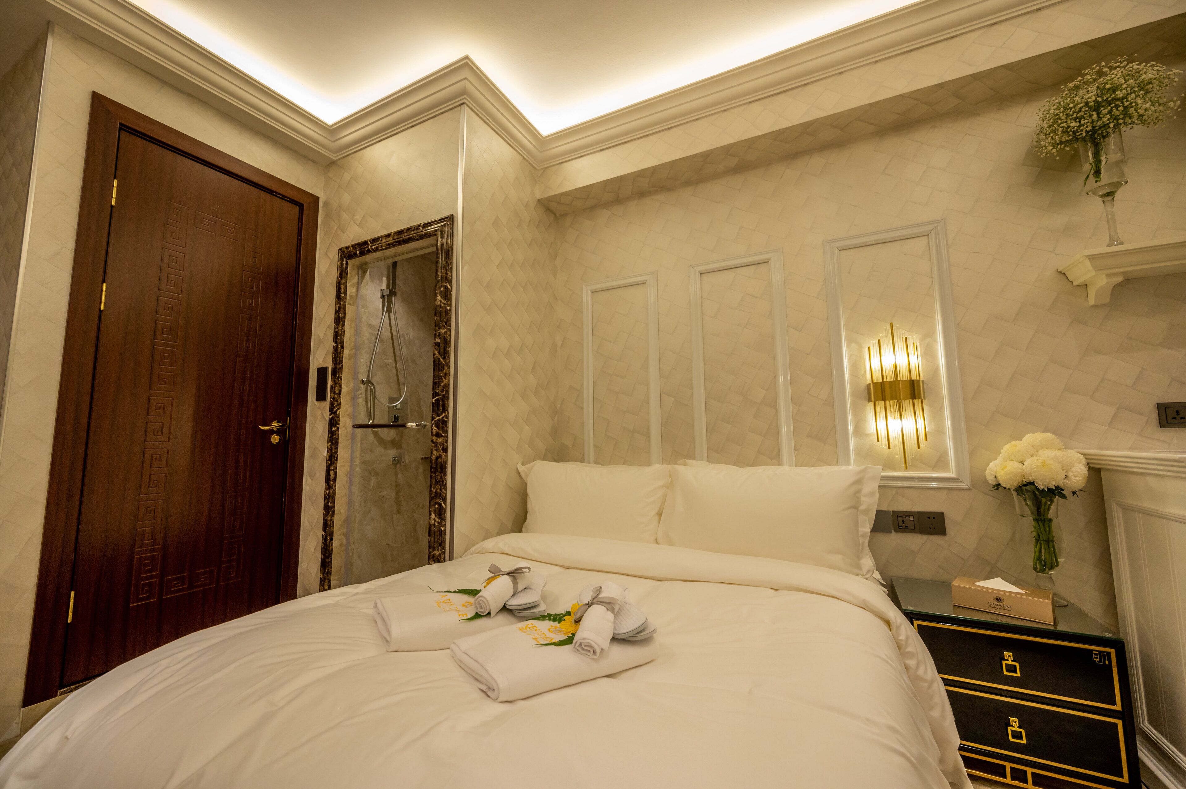 A hotel room with a large bed, a wooden door, a mirror, a nightstand with a vase of flowers, and a wall-mounted light fixture.
