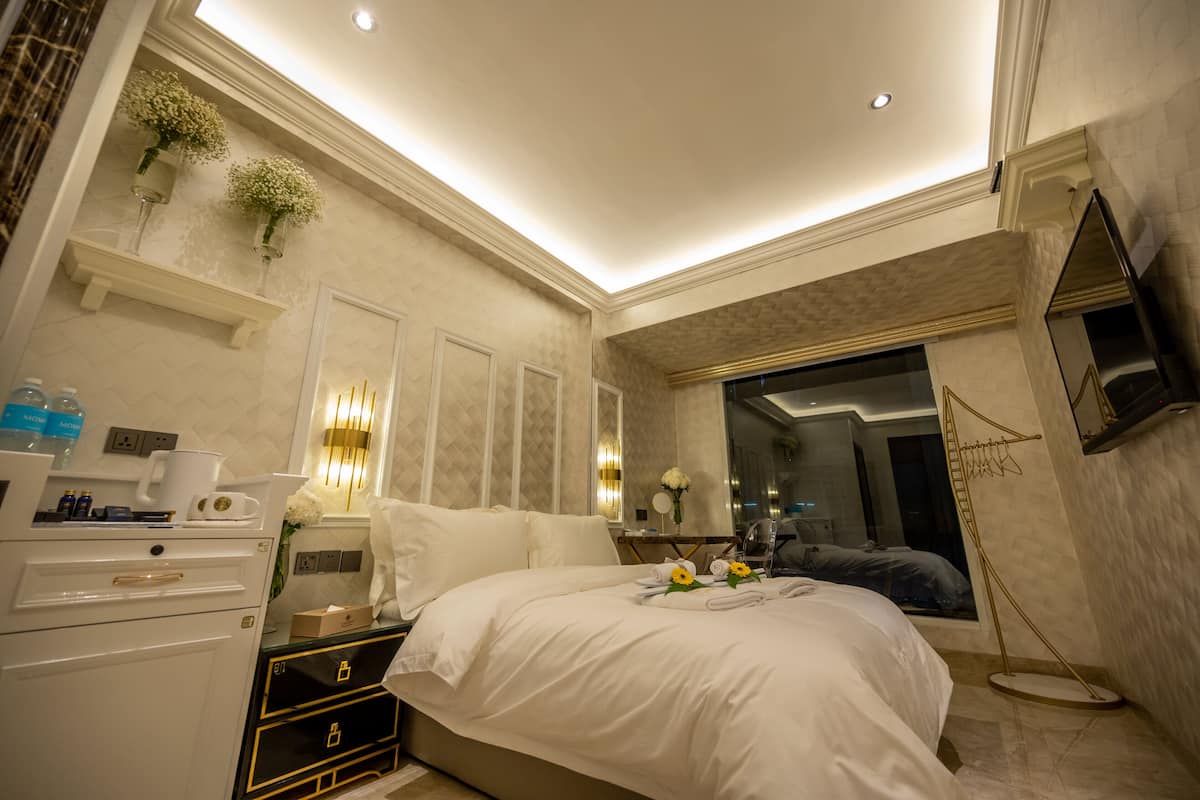 A modern hotel room with a large bed, bedside tables, a vanity, and a bathroom visible through a glass door.