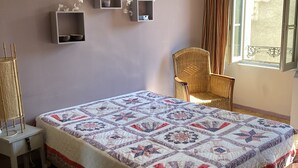 A bedroom with a large bed, a wicker chair, a small table, and wall-mounted shelves. - Large, bright apartment (Argentat-sur-Dordogne)