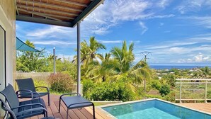 Zona junto a la piscina con un muelle de madera, sillas de descanso y vistas a palmeras y al océano.