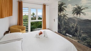 Una habitación de hotel con una cama, una silla, un ventanal con vistas a palmeras y montañas, y un mural de paisaje tropical.