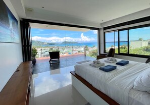 A modern hotel room with a large bed, a seating area, and a balcony with a view of the sea and buildings. - Andaman Hills by Lofty (Patong)