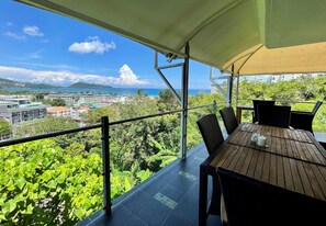 A balcony with a table and chairs, overlooking a lush green landscape and a distant view of buildings and the sea. - Andaman Hills by Lofty (Patong)