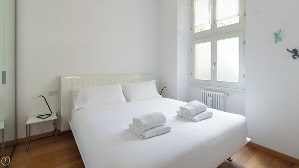 A neatly made bed with white linens and two folded towels, a bedside table with a lamp, and a window with a view of the outside.