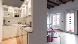 Une cuisine moderne avec des meubles blancs, un plan de travail en granit et un four encastré. On y trouve un coin repas avec une table et des chaises en verre, ainsi qu’une porte donnant sur une pièce lumineuse avec vue sur l’extérieur.