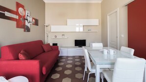 A modern living room with a red sofa, a glass dining table, white chairs, a flat-screen TV, and a kitchen area with white cabinets and a microwave. - Italianway Comfort - Borsieri 11 B (Milan)