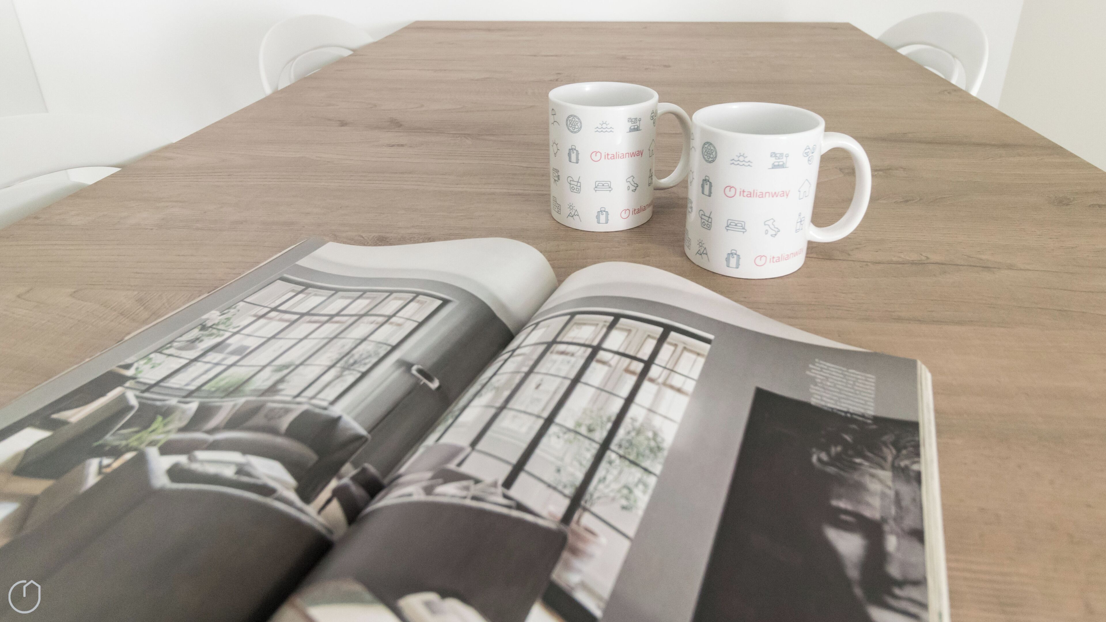 A wooden table with two white mugs featuring Italianway logos, and an open magazine with architectural images.