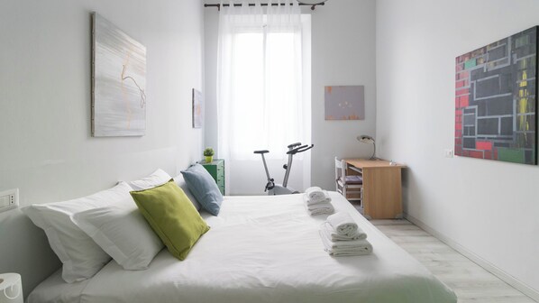 Quarto minimalista com cama, almofadas, escrivaninha e bicicleta de exercício.