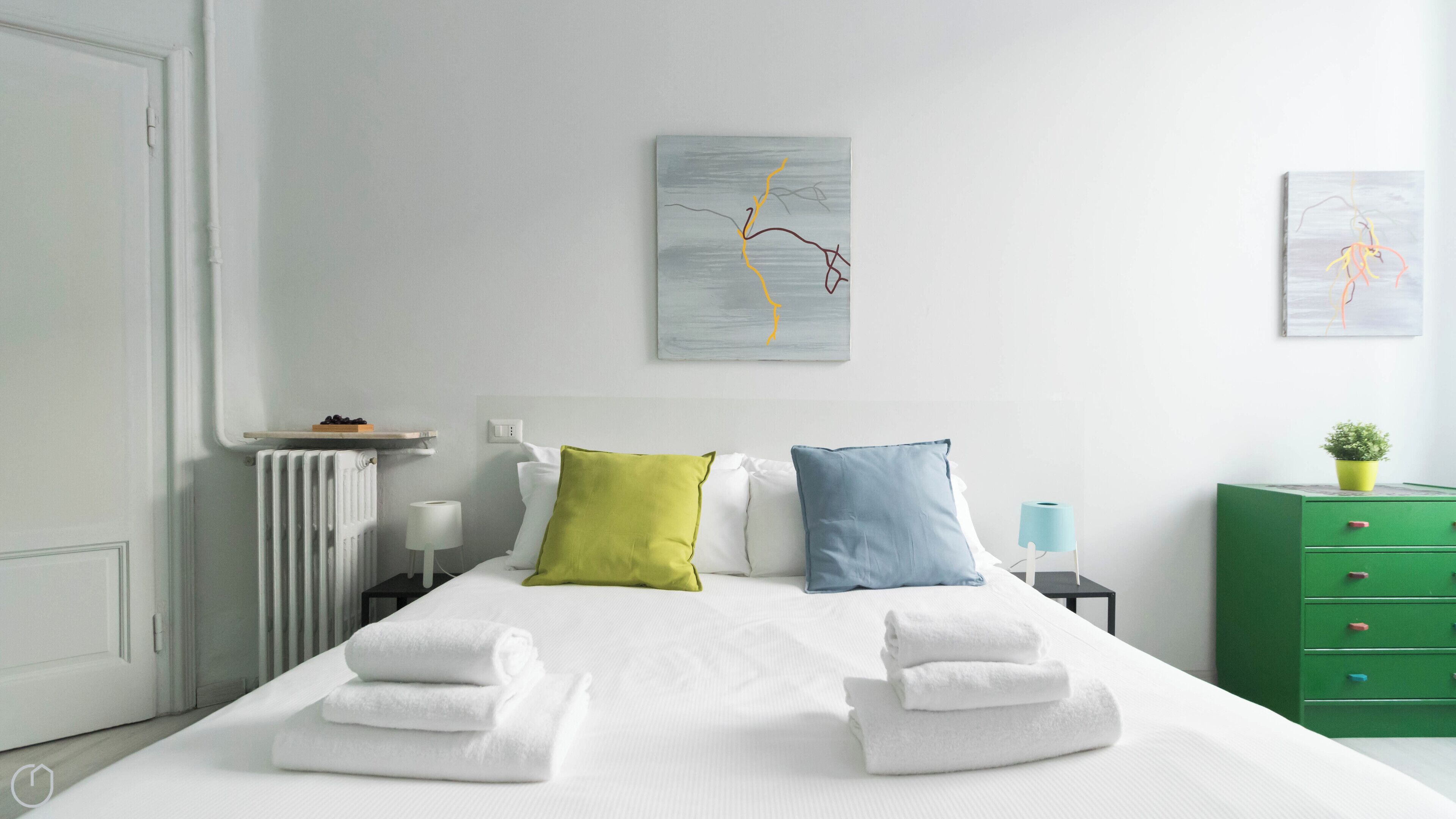 Una cama bien hecha con sábanas blancas y dos almohadas, un aparador verde, una mesita de noche con una lámpara y dos cuadros abstractos en la pared.