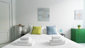 Cama bem arrumada com lençóis brancos e duas almofadas, cômoda verde, mesa de cabeceira com luminária e duas pinturas abstratas na parede.