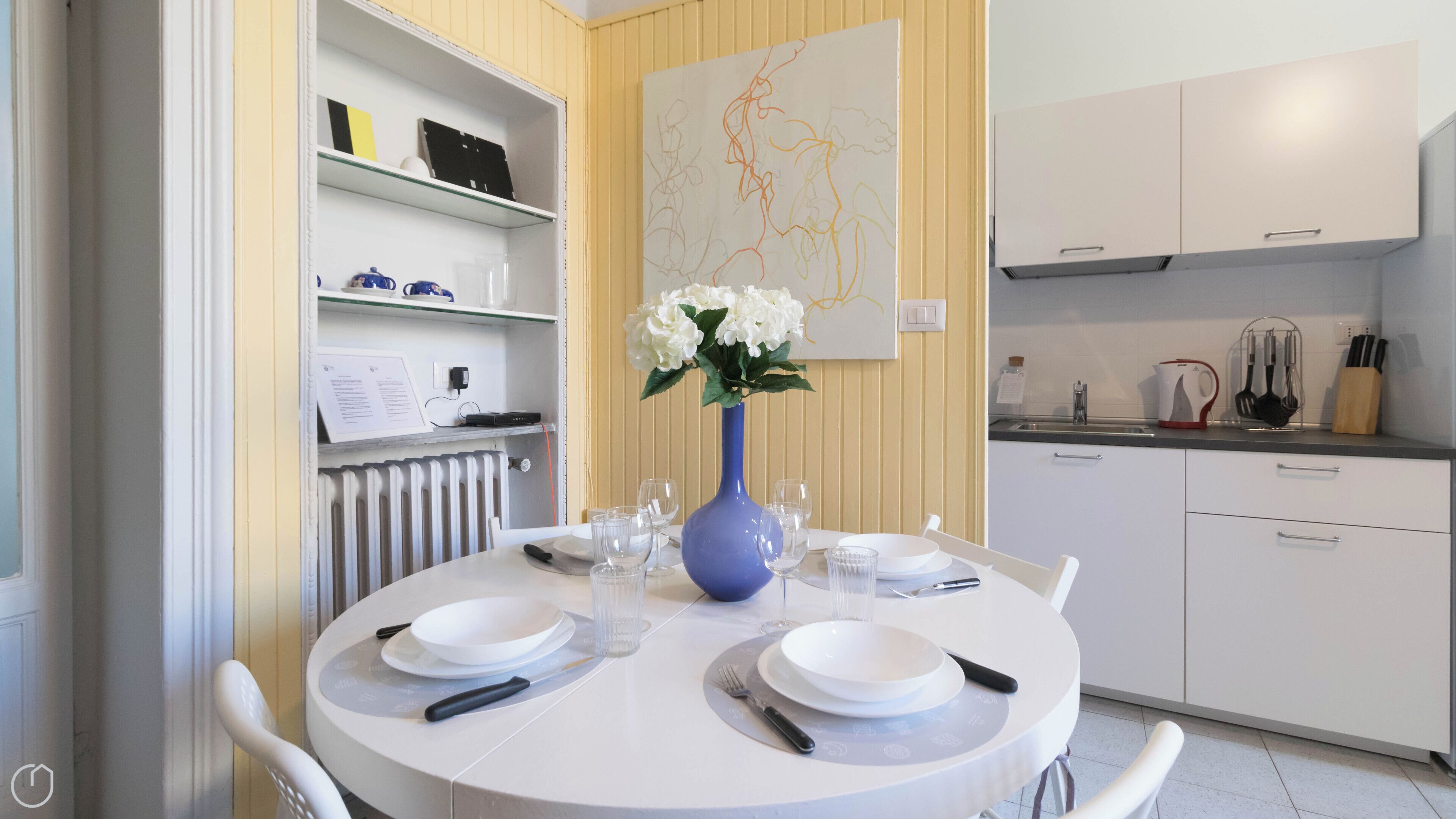 A dining area with a round table set for four, white chairs, a blue vase with white flowers, and a white cabinet with a map on the wall.