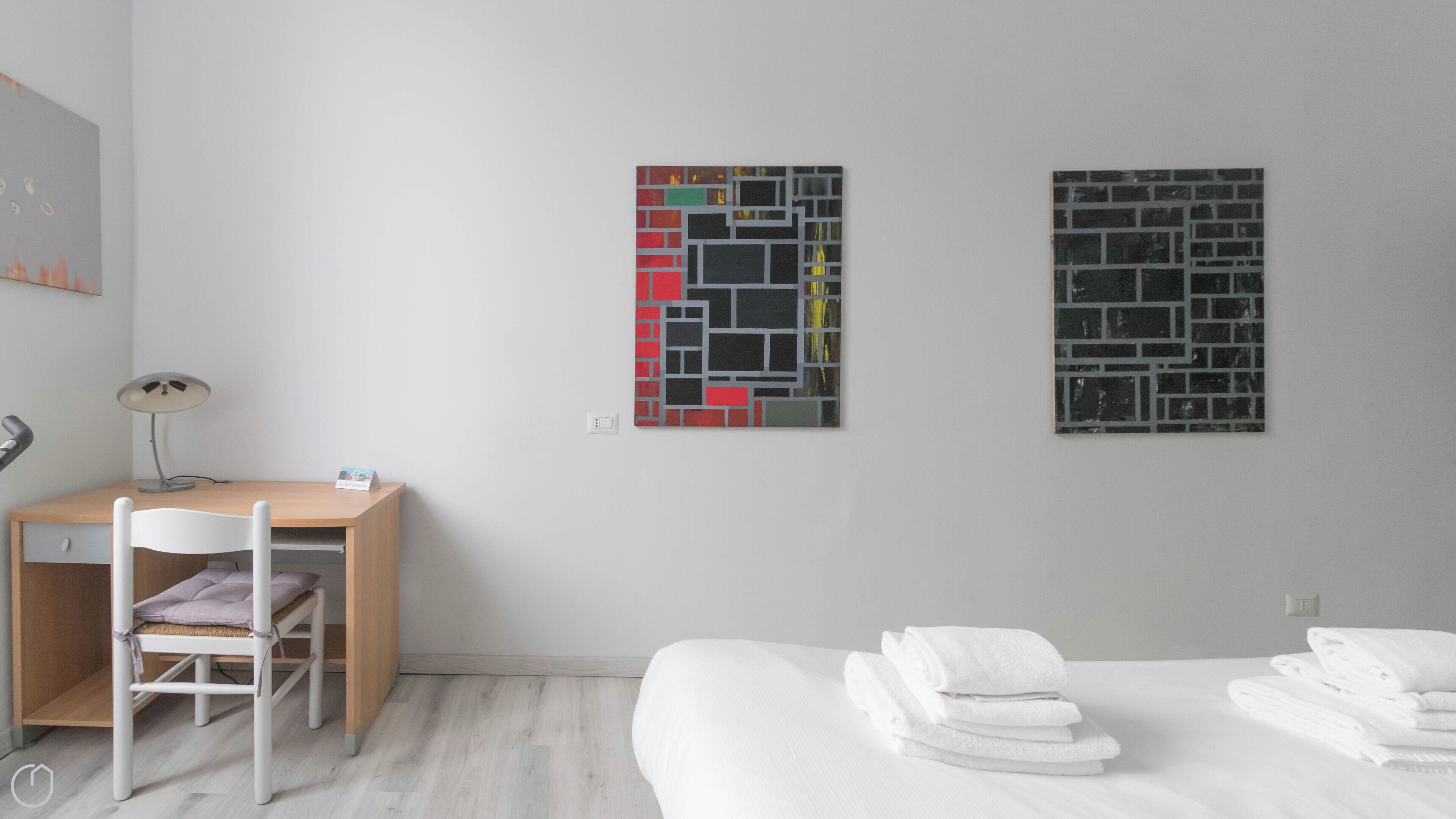 A modern hotel room with a wooden desk, a white chair, a bed with white linens, and two abstract art pieces on the wall.