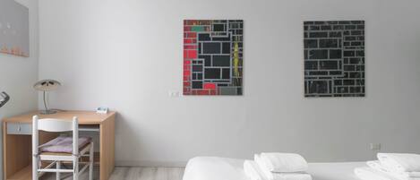 A modern hotel room with a wooden desk, a white chair, a bed with white linens, and two abstract art pieces on the wall.