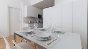 Un comedor moderno con una mesa dispuesta para cuatro, sillas blancas y una cocina al fondo.
