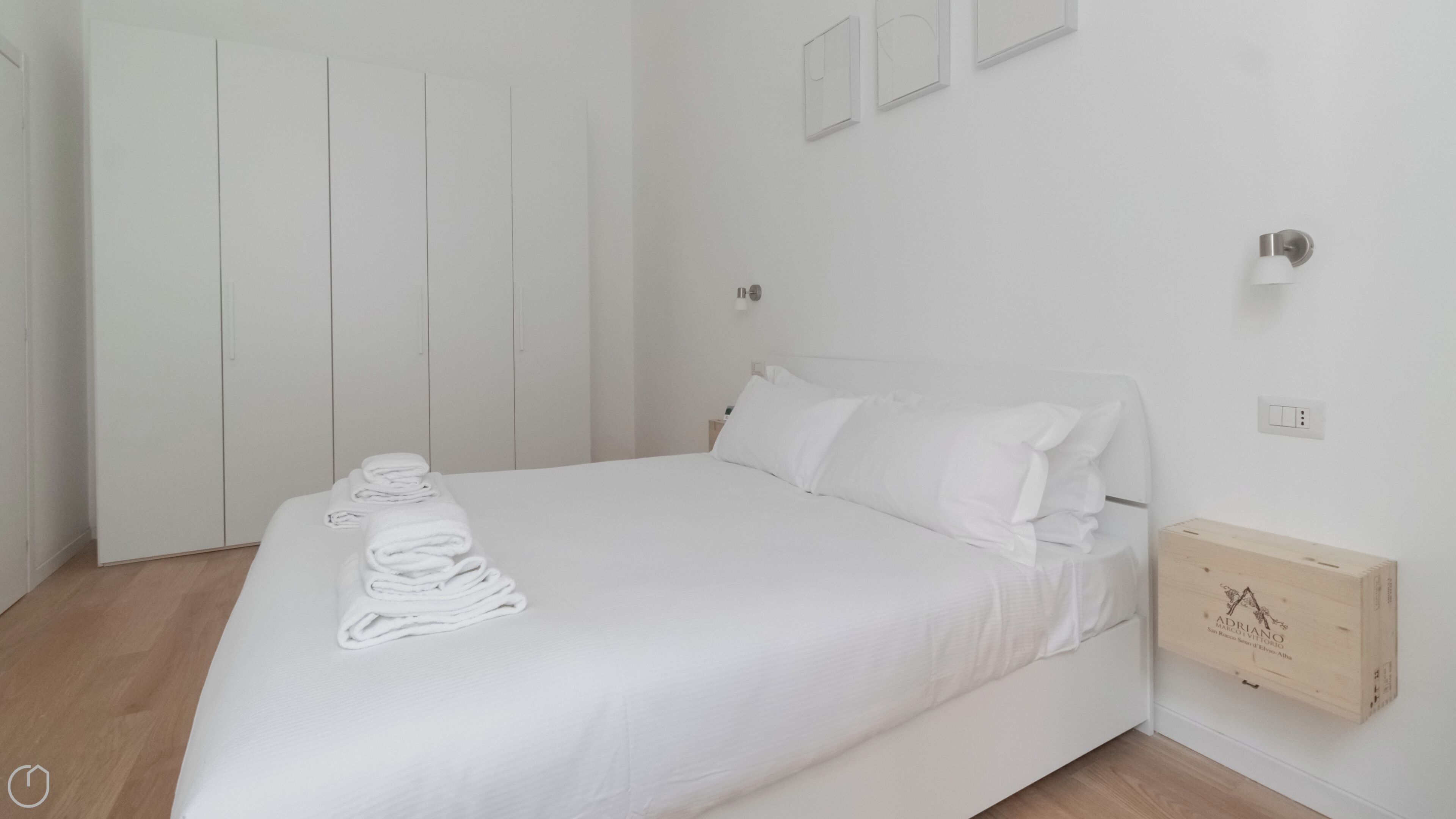 A neatly made bed with white linens, a wooden nightstand with a lamp, and a wooden wardrobe.