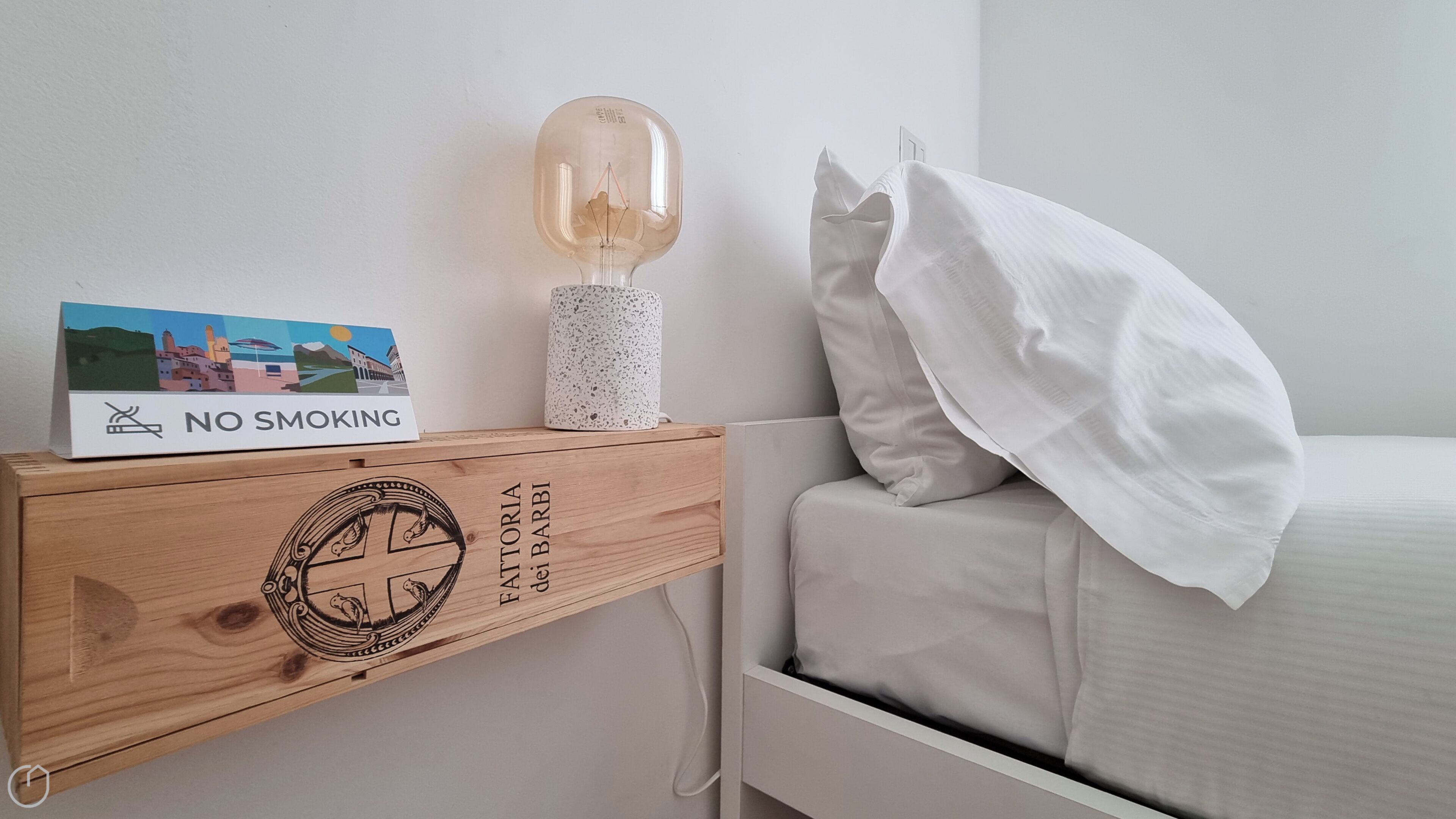 A bedroom with a wooden nightstand, a 'No Smoking' sign, a decorative lamp, and a bed with a white pillow.