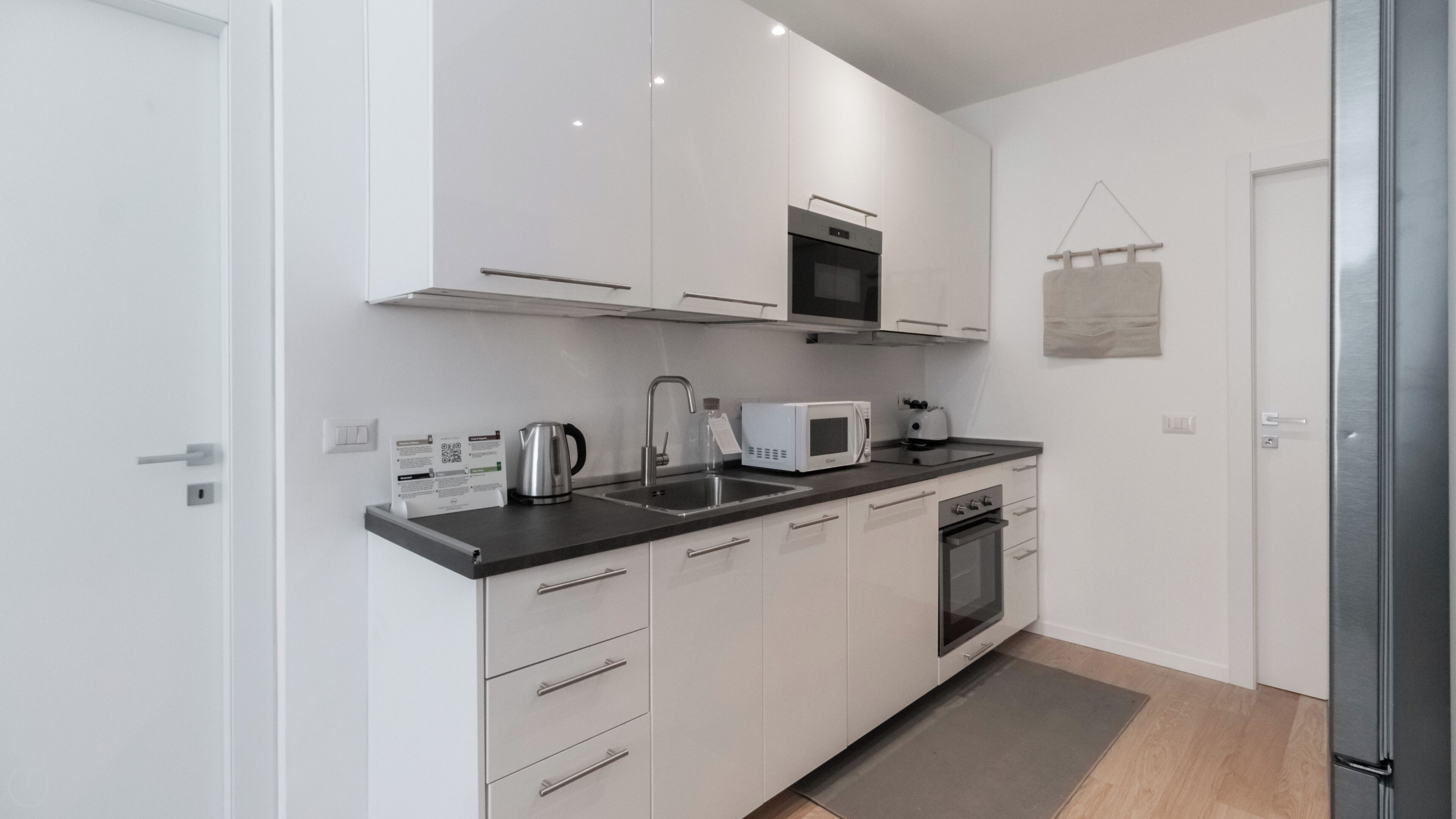 A modern kitchen with white cabinets, a stainless steel sink, a microwave, a kettle, and an oven.