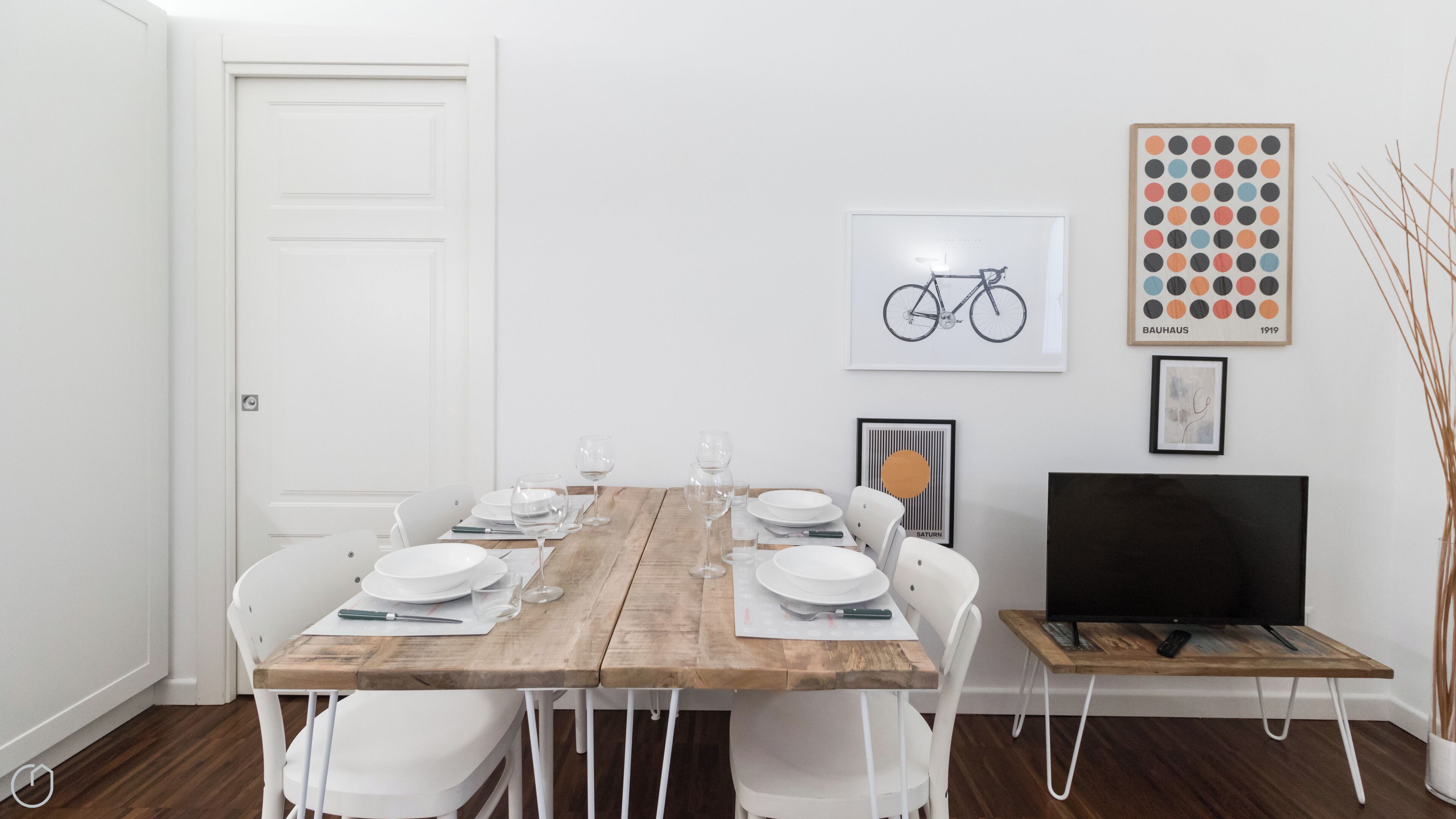 A dining area with a wooden table set for four, white chairs, a white door, and a television on a stand.