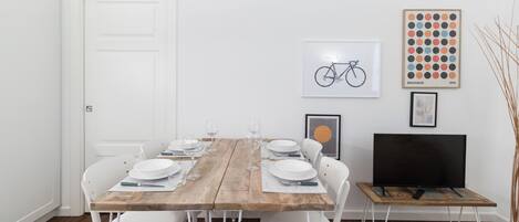 A dining area with a wooden table set for four, white chairs, a white door, and a television on a stand.