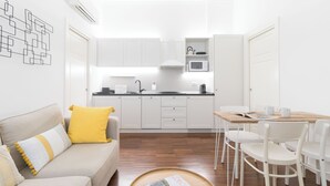 A compact kitchen with white cabinets, a microwave, and a sink. There is a dining area with a wooden table and white chairs. A sofa with yellow and white cushions is in the foreground. - Italianway - Jacopo dal Verme 4 (Milan)