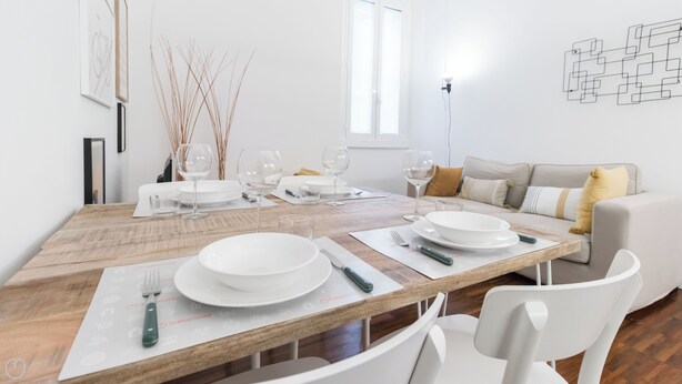 A dining area with a wooden table set for four, white chairs, and a sofa in the background. - Italianway - Jacopo dal Verme 4 (Milan)