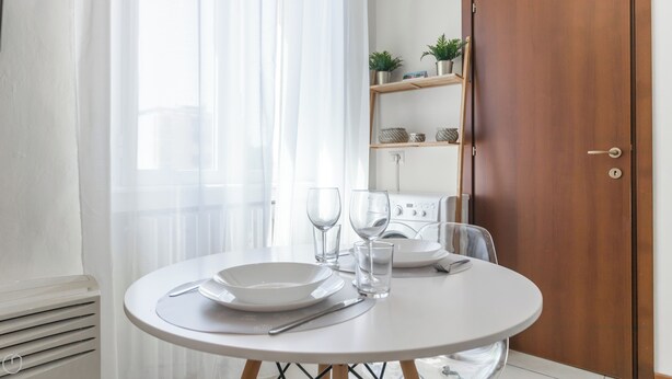 A round dining table with white plates, cutlery, and glasses, set against a white wall with a wooden door and a shelf with decorative items. - Italianway - Gallura 9 (Milan)