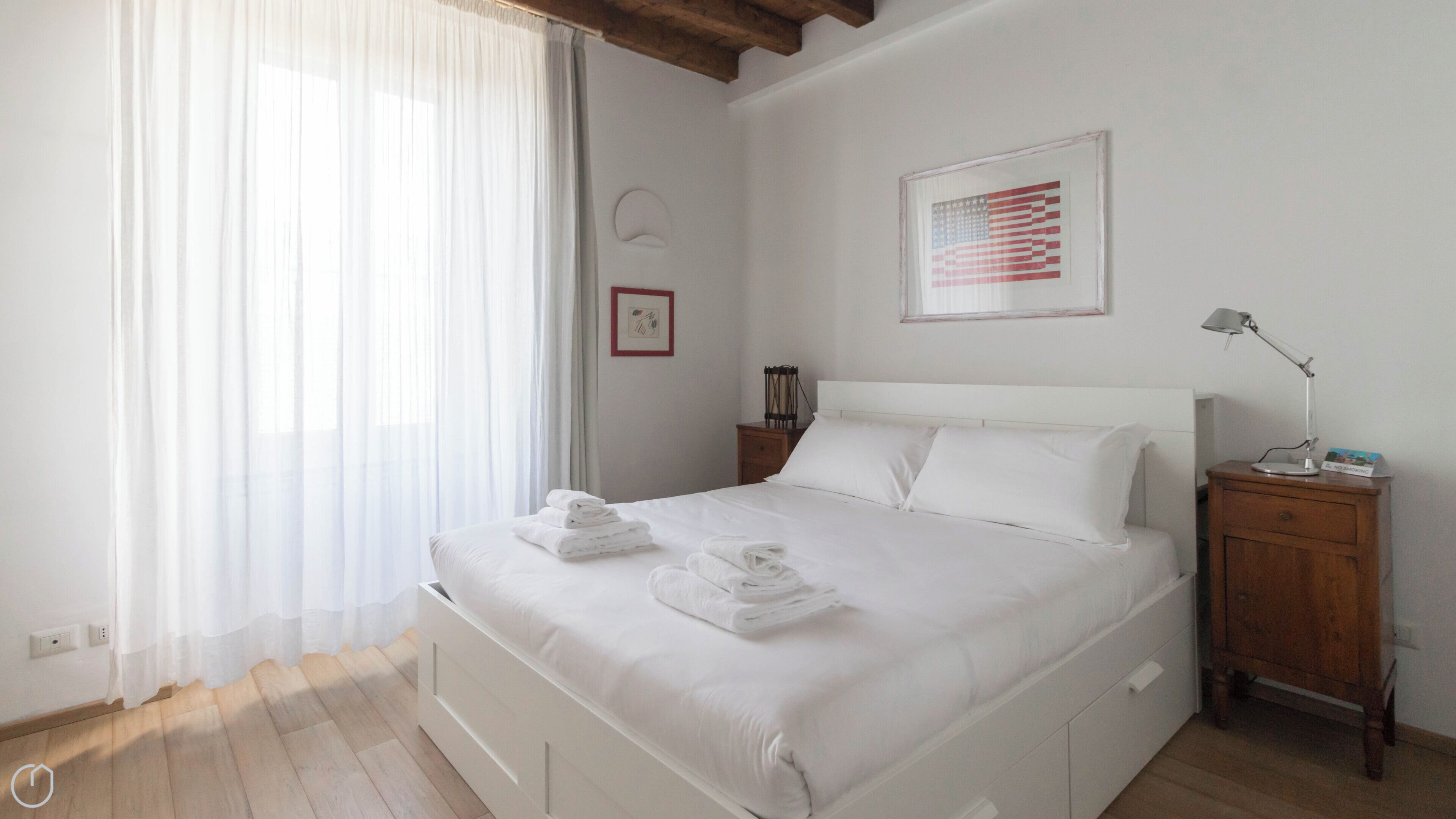 A neatly made bed with folded towels, a bedside table with a lamp, and a framed American flag on the wall.