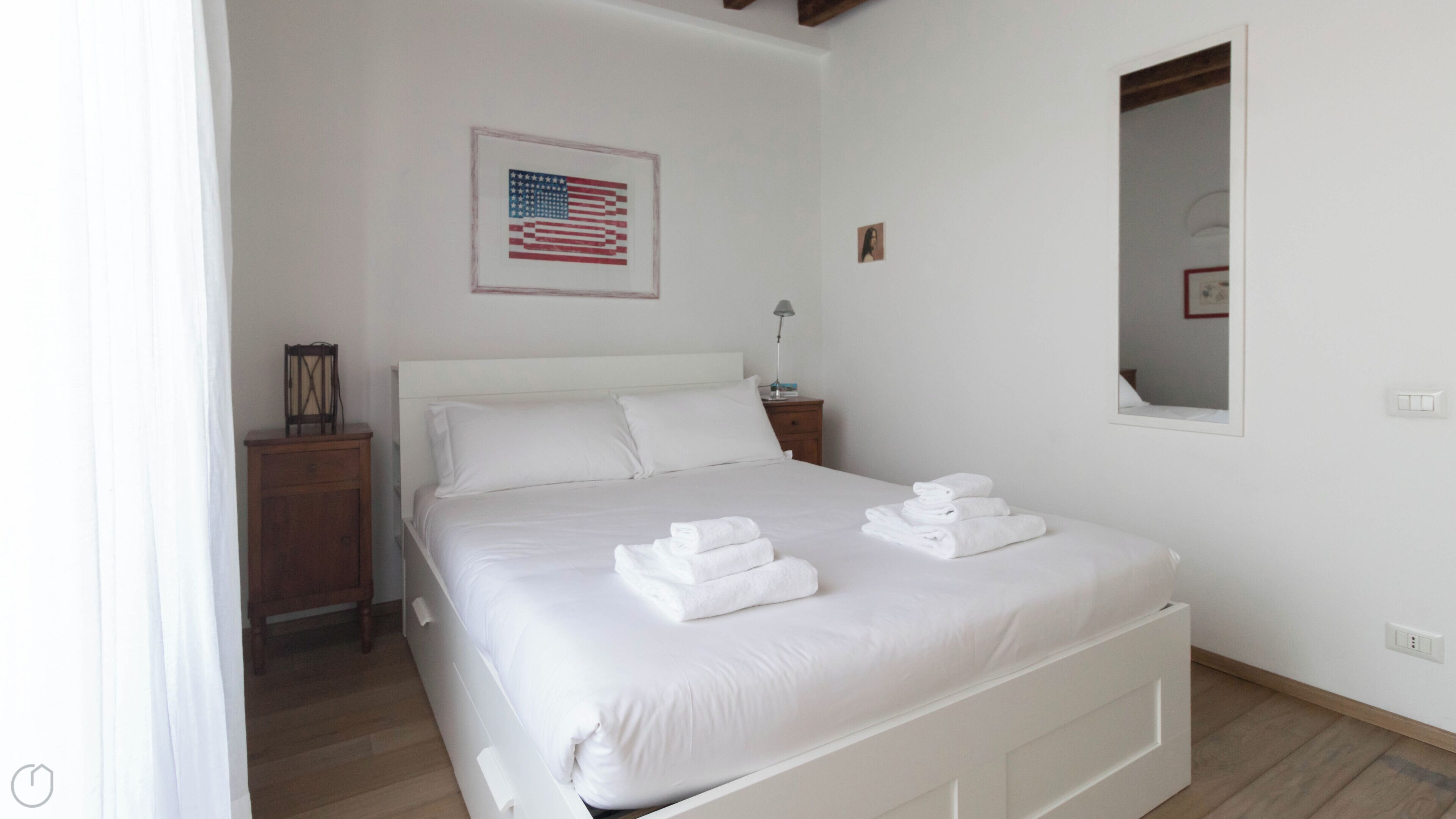 Ein ordentlich bezogenes Bett mit weißen Bettwäsche und Handtüchern, ein Nachttisch aus Holz mit einer Lampe und eine gerahmte amerikanische Flagge an der Wand.