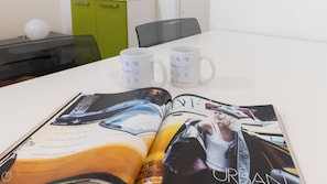 A magazine spread on a table with two white mugs with Italianway logos, a green cabinet, and a black chair in the background. - Italianway Easy - Plinio 64 (Milan)