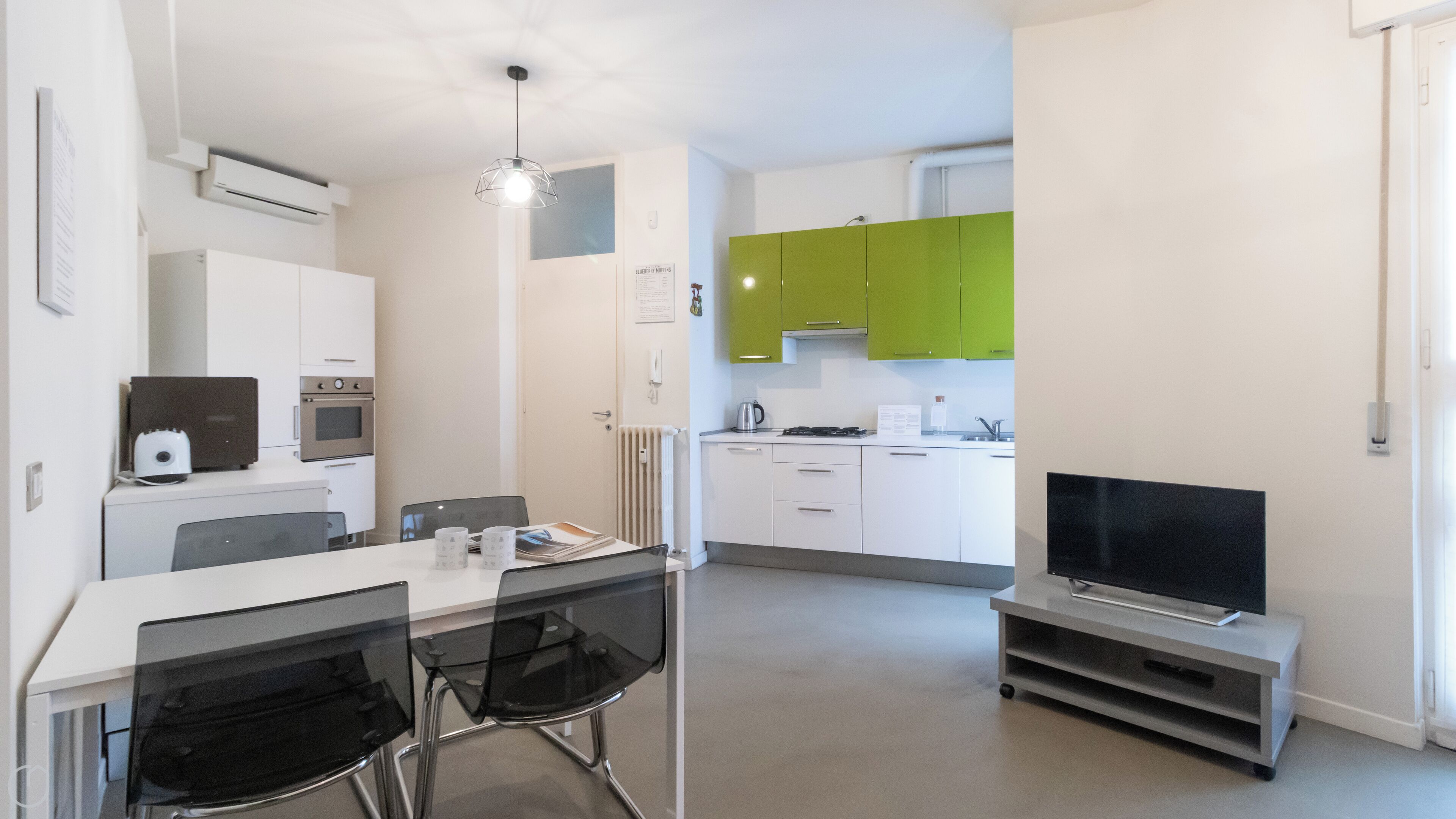 A modern kitchen with white and green cabinets, a dining area with a table and chairs, and a flat-screen TV on a stand.