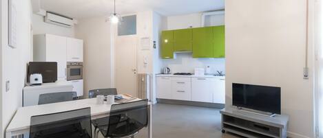 A modern kitchen with white and green cabinets, a dining area with a table and chairs, and a flat-screen TV on a stand.