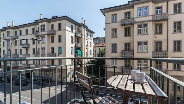 A balcony with a wooden table and chairs, overlooking a street with parked cars and buildings. - Italianway Easy - Sanfelice 10 (Milan)