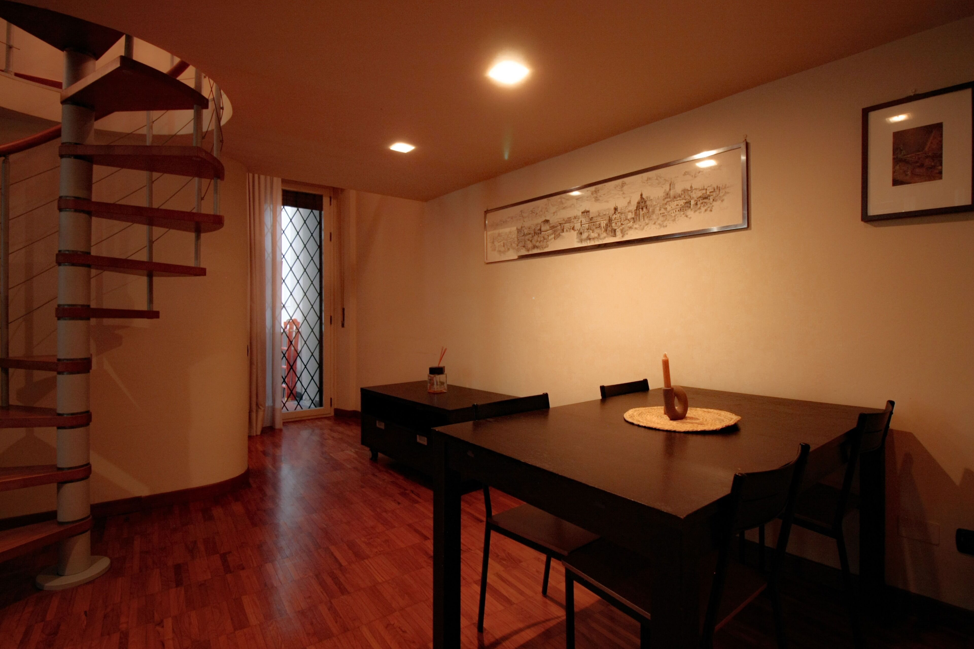 Un coin repas avec une table et des chaises en bois, un escalier en colimaçon et un tableau encadré au mur.