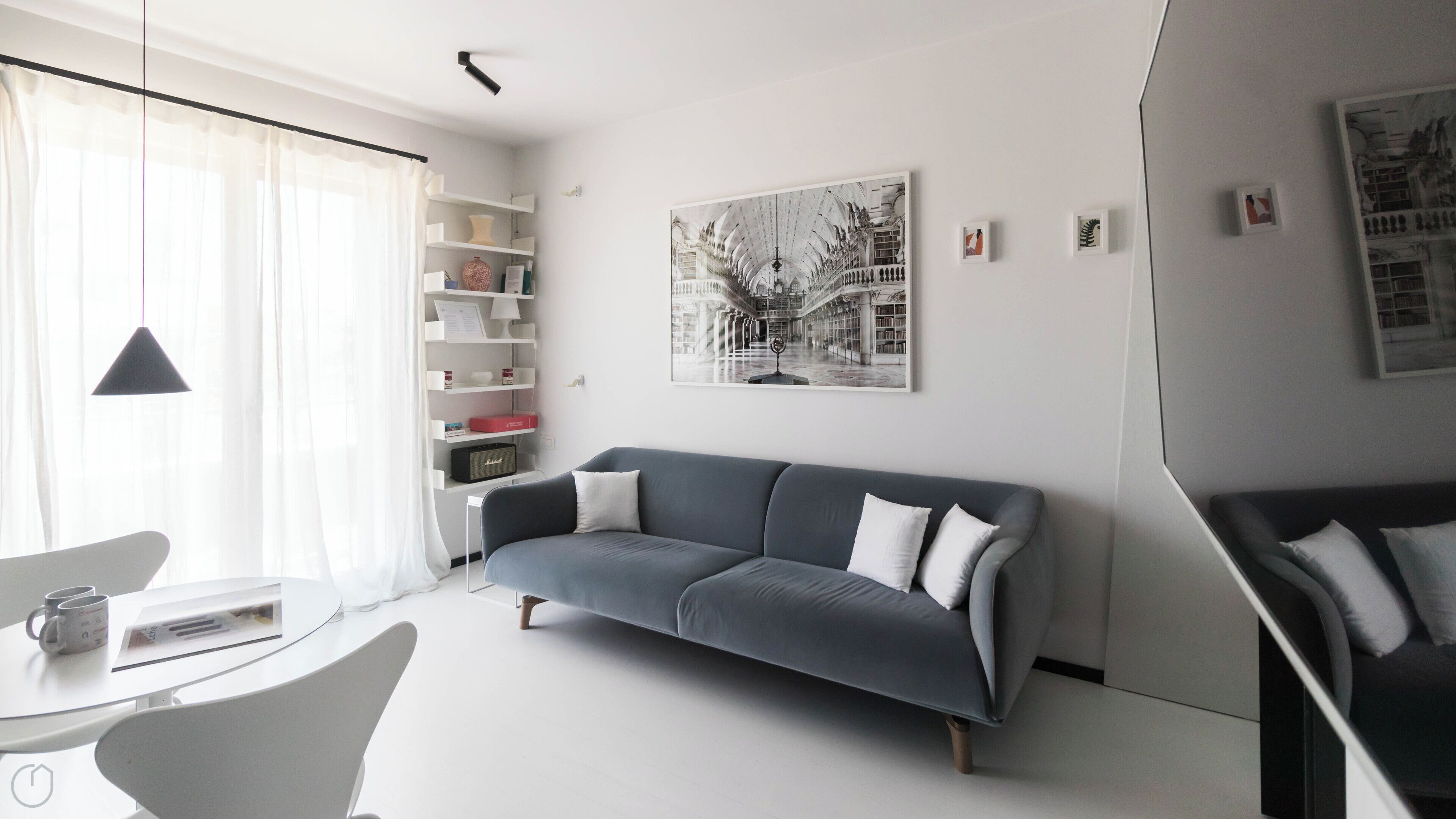 Una sala de estar moderna con un sofá gris, cojines blancos, una mesa de centro blanca y una lámpara negra. Se observa una estantería con varios objetos y un cuadro enmarcado en la pared.