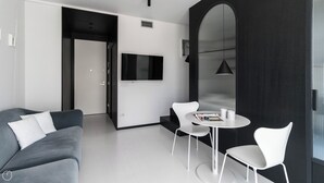 Un salon moderne avec un canapé gris, des fauteuils blancs, une petite table ronde et un téléviseur fixé au mur.
