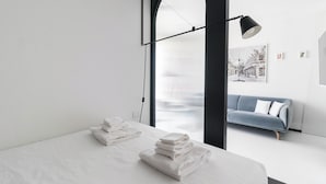 Une chambre d’hôtel moderne avec un lit, une pile de serviettes blanches, un canapé bleu et une grande fenêtre donnant sur un immeuble.