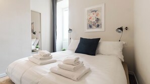 Ein ordentlich bezogenes Bett mit weißen Leinen und gefalteten Handtüchern, ein dunkelblaues Kopfkissen und ein Gemälde an der Wand.