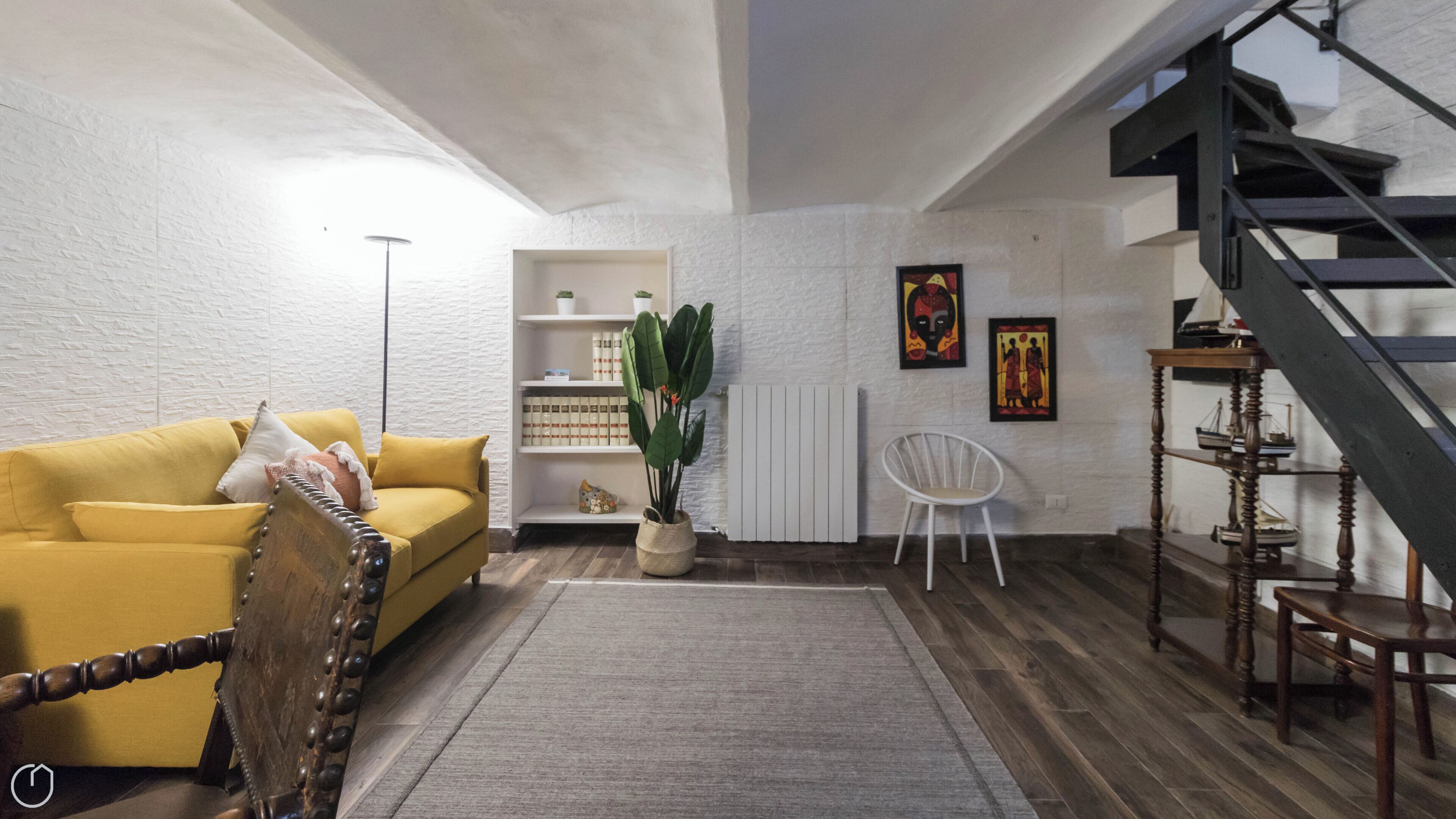 Ein Wohnzimmer mit gelber Couch, weißem Sessel, Bücherregal, Pflanze und Treppe.