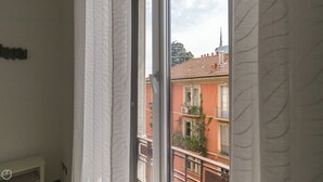 Ein Fenster mit weißen Vorhängen, Blick auf ein Gebäude mit roten Wänden und einem Ziegeldach, sowie ein Balkon mit Geländern.