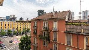 Ein mehrstöckiges Gebäude mit Balkonen, eine Straßenszene mit Autos und Fußgängern sowie moderne Hochhäuser im Hintergrund.