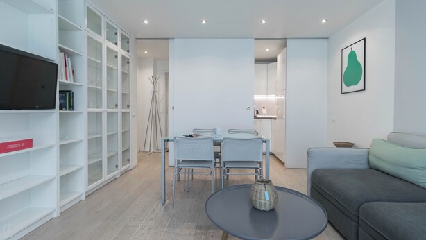 A modern living room with a grey sofa, a round coffee table, and a dining area with a glass table and metal chairs. There is a bookshelf with books and a framed picture on the wall.