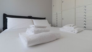 A neatly made bed with white linens and towels, a black headboard, and a white wardrobe with silver handles.