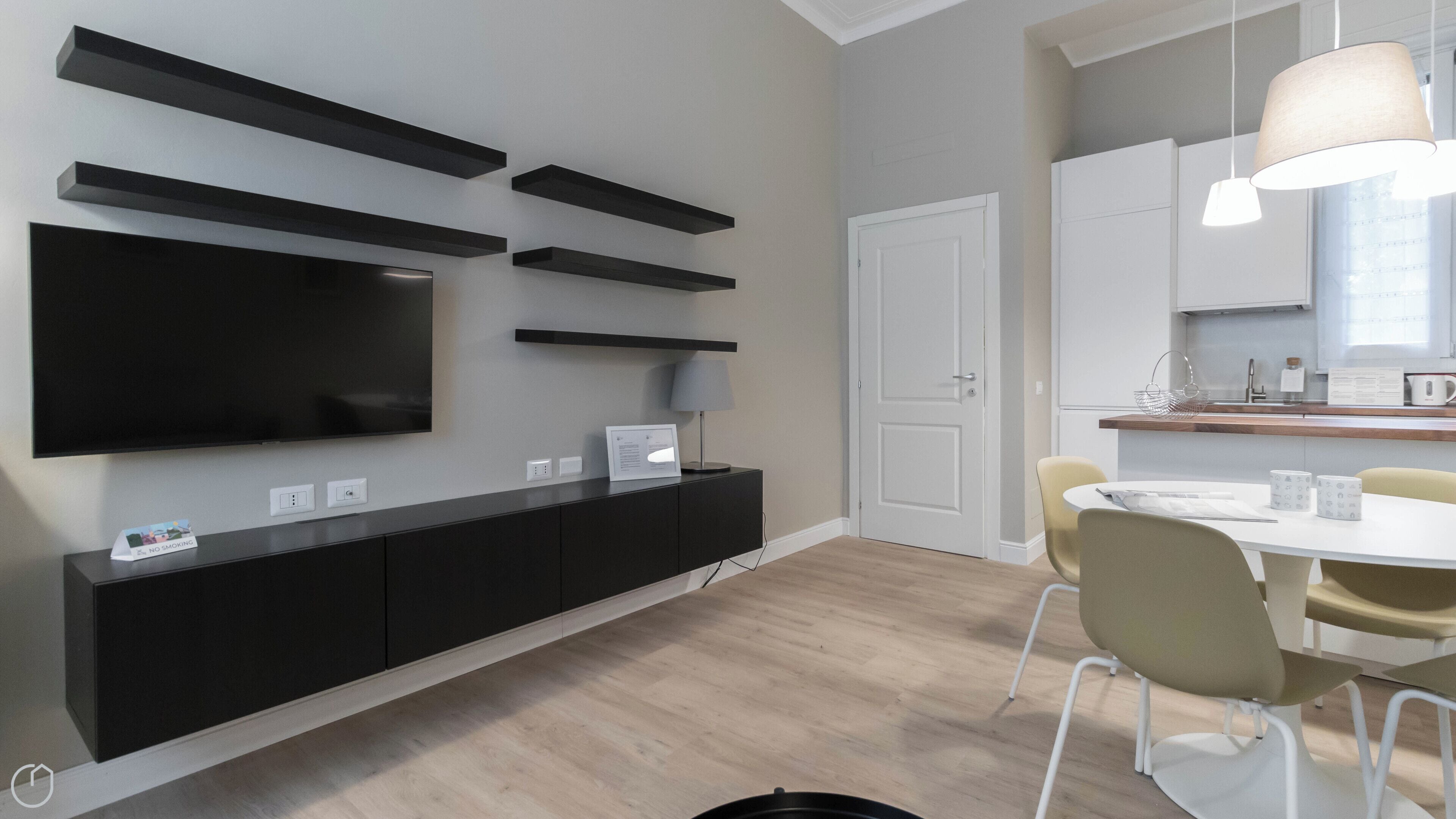 A modern kitchen with a built-in oven, a dining table with chairs, and a large flat-screen TV mounted on the wall.